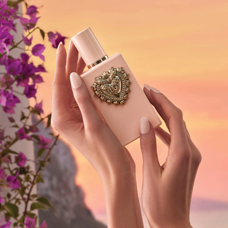 Hand holding a pink perfume bottle with decorative cap against a blurred floral background