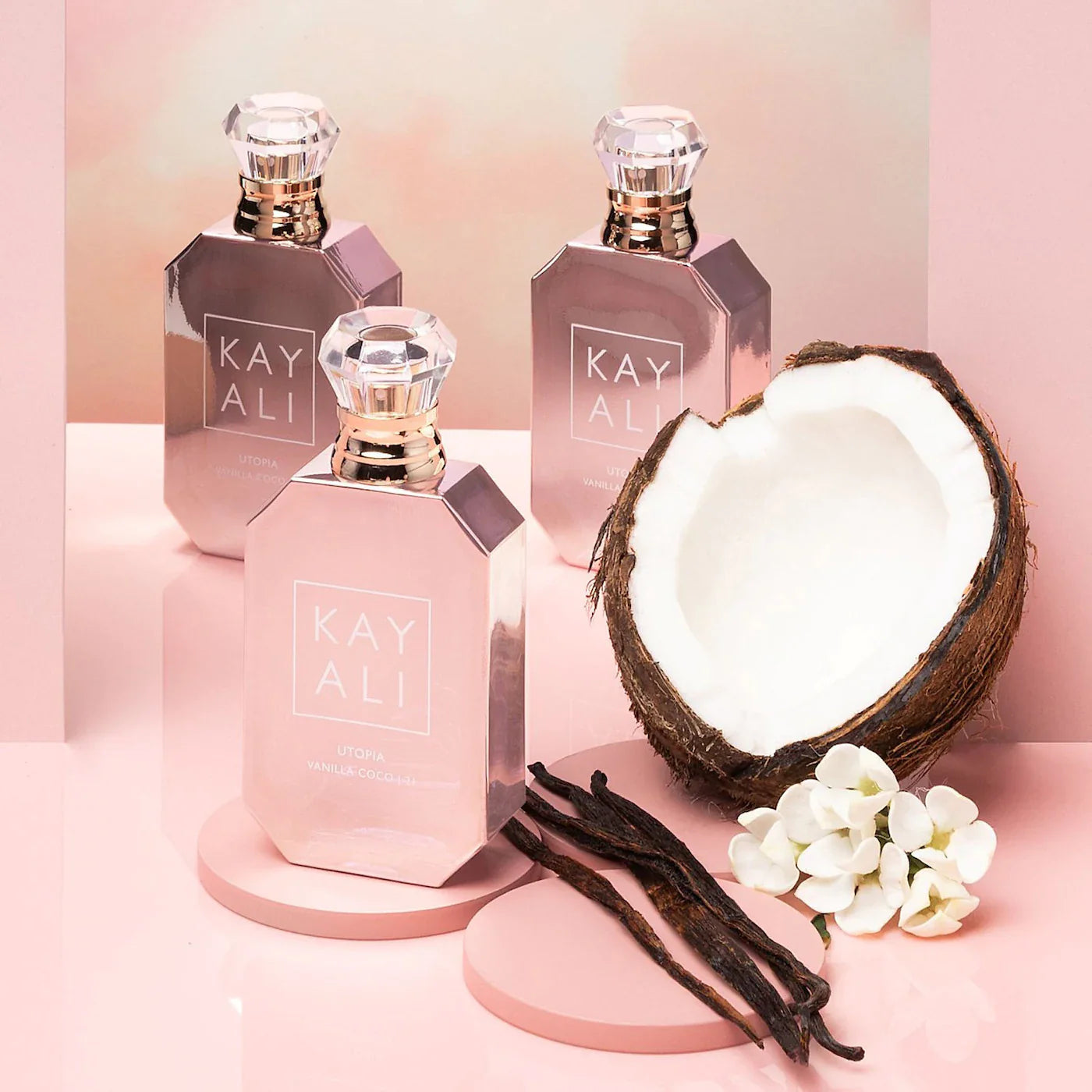Three 'KAY ALI' perfume bottles on a pink background with a coconut, vanilla beans, and flowers.